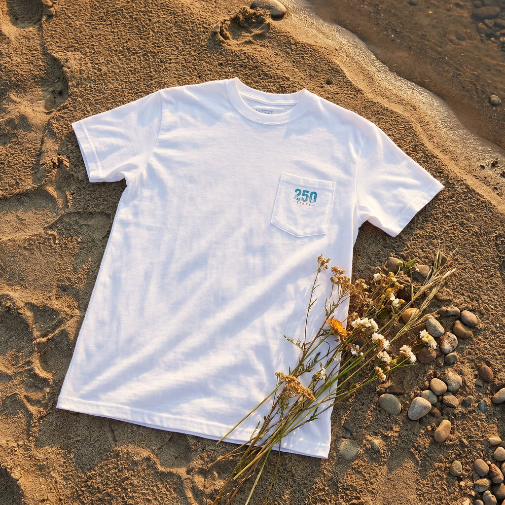 America 250th Anniversary Tee flat-lay on sandy riverbank with wildflowers