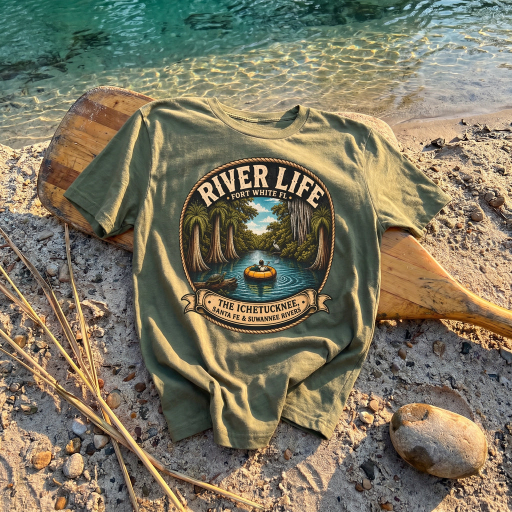 River Life Mens Tee — flat-lay on riverbank with canoe paddle, teal spring water, river reeds