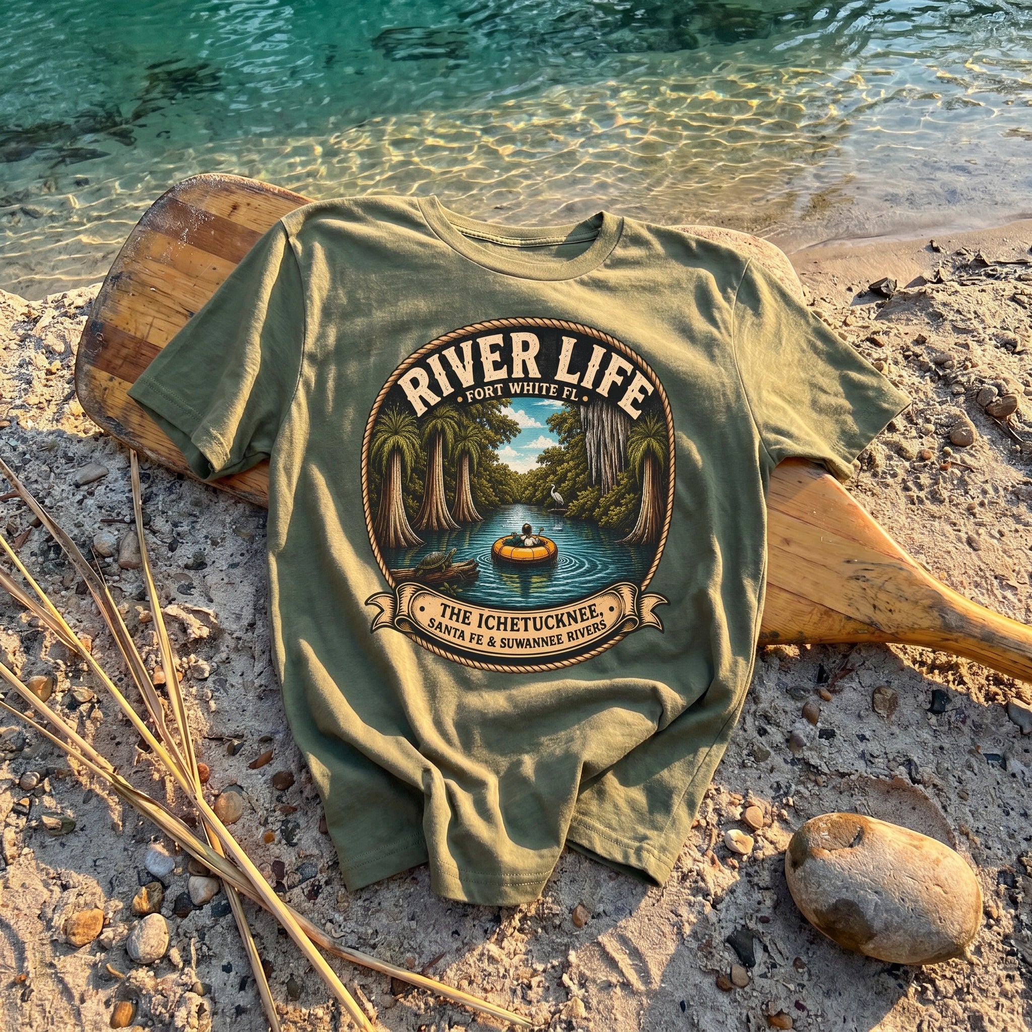 River Life Mens Tee — flat-lay on riverbank with canoe paddle, teal spring water, river reeds